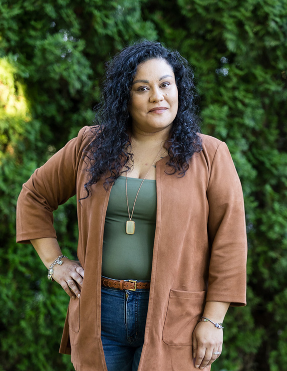 hispanic professional business woman posing outside in a green top, jeans, and brown jacket