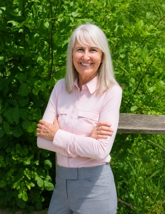 paula-2026 white middle-aged business woman posing for staff photo in pink blouse and grey slacks - platinum grey hair and green bush in background